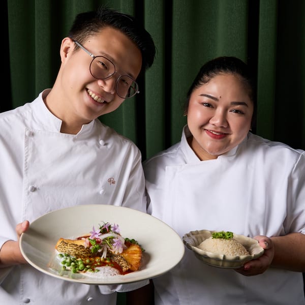 Two smiling Khaosan chefs in white coats hold a salmon dish and a bowl of rice.