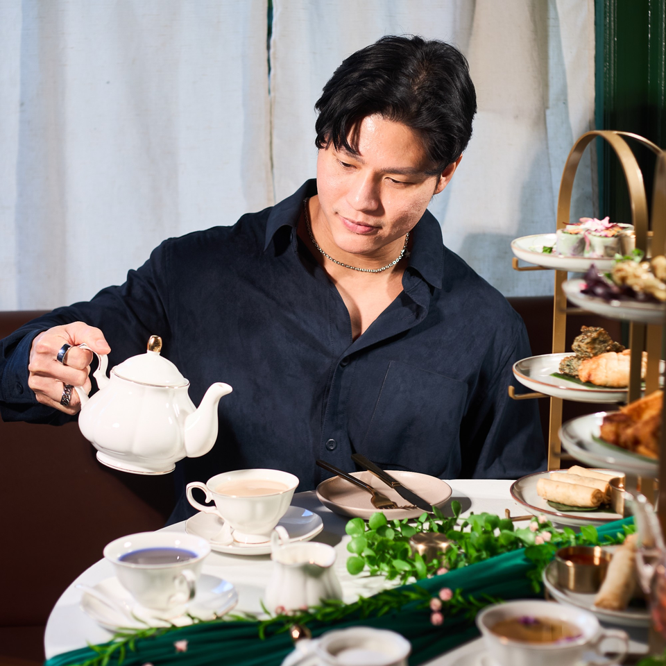 A man pours tea from a white teapot into a cup at a table with Thai appetizers.
