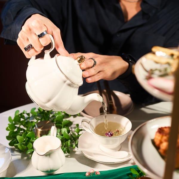 A person pouring hot tea from a white teapot into a teacup with dried flowers.