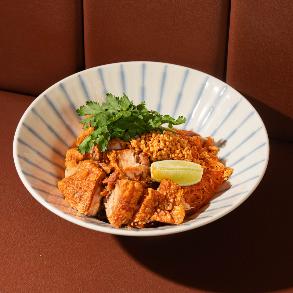Thai-style vermicelli bowl topped with crispy pork, minced peanuts, cilantro, and a lime wedge on a striped ceramic plate.