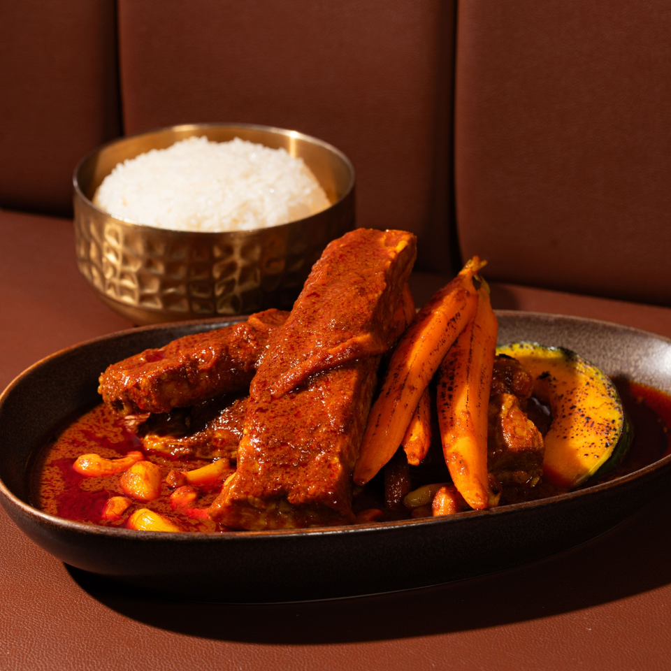 Spiced braised meat with roasted carrots and squash in red sauce, served with white rice in a bowl.