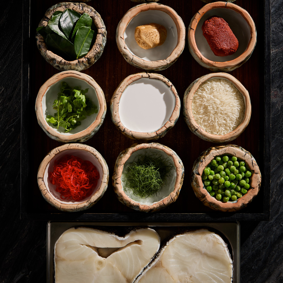 Nine bowls of Thai ingredients, including herbs, spices, rice, and peas, with raw fish below.