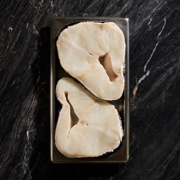 Two raw, thick-cut fish fillets in a metal tray on a dark marble surface.