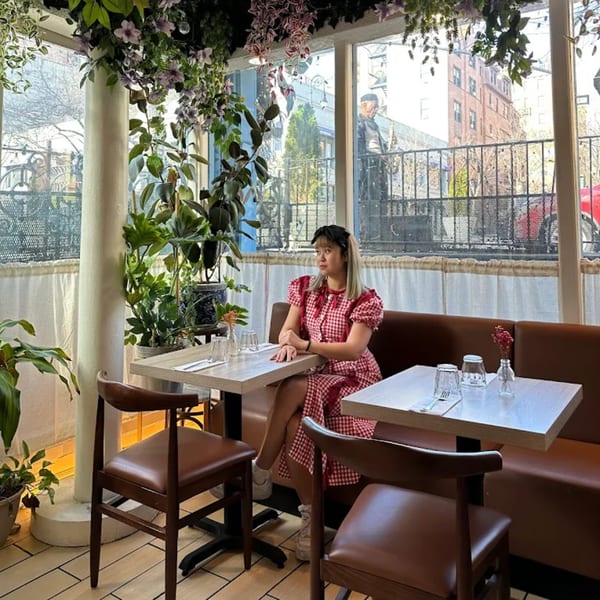 A woman in a red gingham dress sits at a table in a bright, plant-filled Khaosan restaurant.