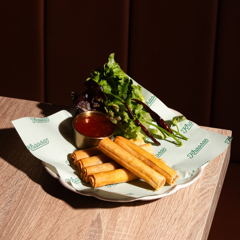 Crispy spring rolls with fresh greens, dried chilies, and sweet chili dipping sauce on a branded plate.