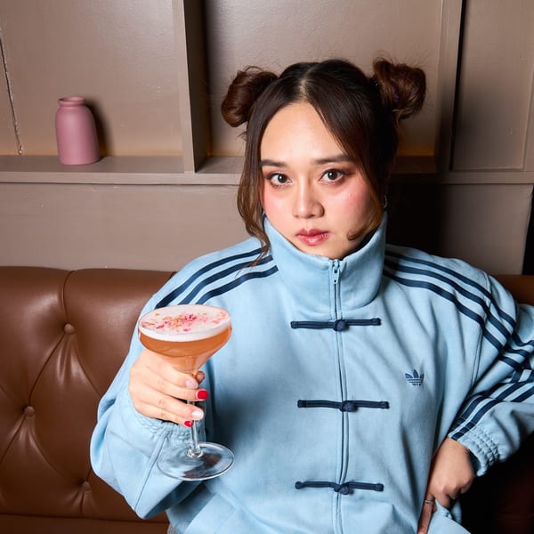 A woman in a blue jacket holds a pink cocktail with foam and flower petals on a brown couch.